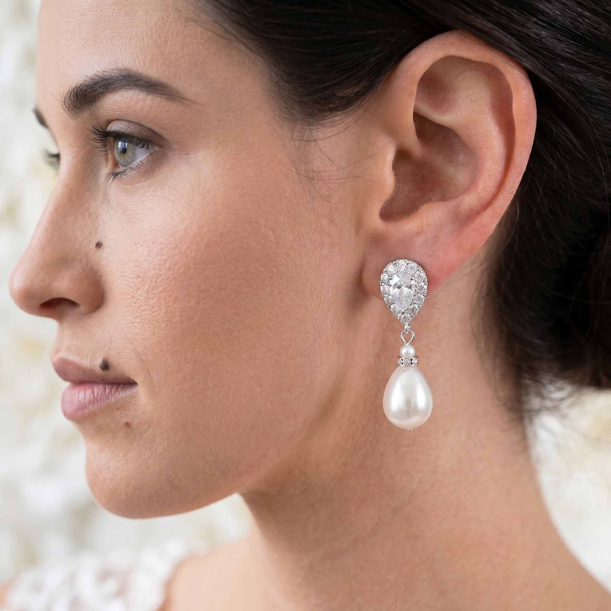 A close-up side profile of a woman wearing a dangling pearl earring with a sparkling silver-toned teardrop-shaped setting.