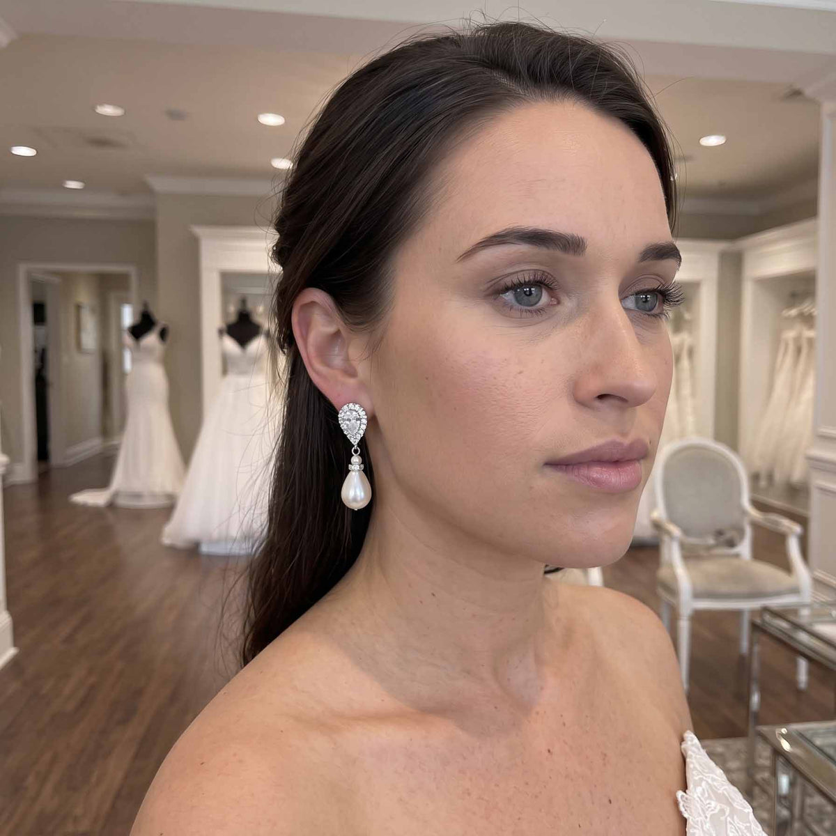 A Bride / woman in a bridal salon wearing a dangling pearl earring with clear gemstone detailing, with wedding dresses visible in the background.