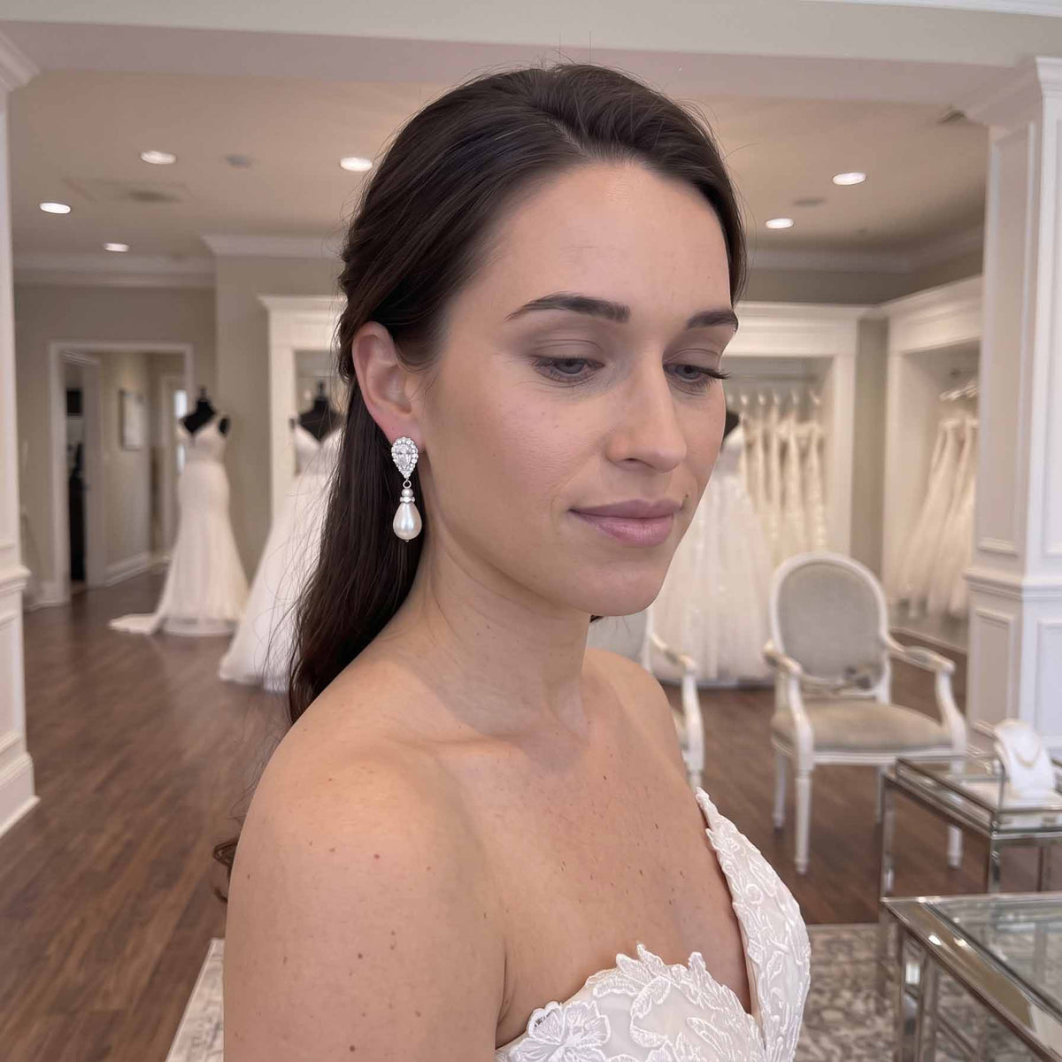 A bride in a white lace bridal gown wearing a silver teardrop-shaped drop earring with clear gemstones, photographed inside a bridal salon with wedding dresses visible in the background.