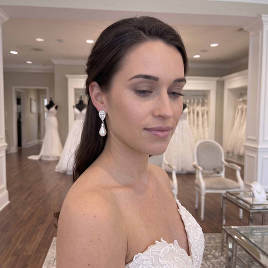 A bride in a white lace bridal gown wearing a silver teardrop-shaped drop earring with clear gemstones, photographed inside a bridal salon with wedding dresses visible in the background.